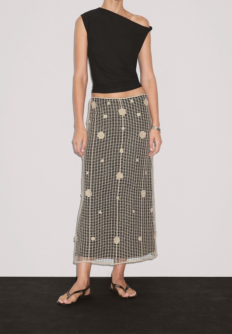 Haut noir à une épaule associé à une jupe longue, à motifs, semi-transparente, présentant un design à carreaux et des accents circulaires de couleur beige.