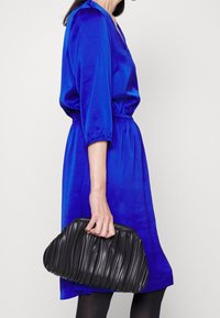 Bolso de mano negro de canalé sostenido por una persona que lleva un vestido de satén azul con cintura elástica y mangas tres cuartos.