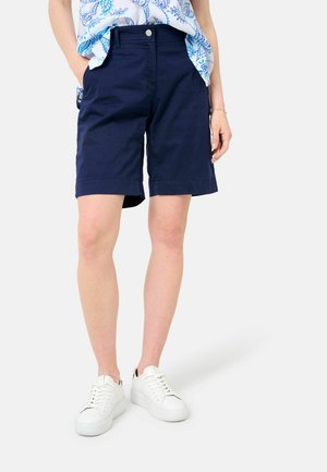 Femme portant un short bleu marine longueur genoux, des baskets blanches et un haut bleu à motifs, les mains dans les poches, sur un fond uni.