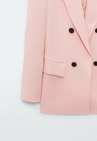 Blazer rose clair à double boutonnage avec boutons noirs, col à revers cranté et poche à rabat à l'avant, présenté sur un fond blanc uni.