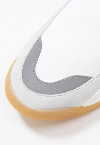 Baskets blanches avec accents en suède gris, design incurvé et semelle extérieure en caoutchouc gomme. Présente un tissu lisse et des détails de couture.