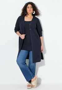 Cardigan long marine avec trois boutons, associé à un débardeur marine, un jean bleu évasé et des sandales plates crème avec des accents floraux.