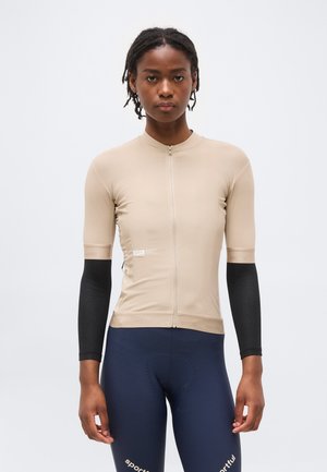 Naine, kes kannab beežikat lühikeste varrukatega rattasärki, musti käesoojendajaid ja tumesiniseid tekstuuriga rattapükse logodega.