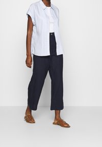 Ljusblå randig kortärmad skjorta över en vit topp, i kombination med löst sittande marinblå byxor och bruna sandaler med remaccent.