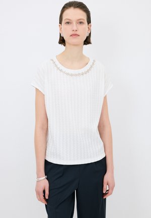 Débardeur blanc à manches courtes avec un motif texturé, encolure ornée de petites perles, associé à un pantalon bleu marine.