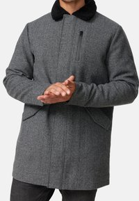 Abrigo gris de espiga con un cuello de piel sintética negro, bolsillo delantero con cremallera y bolsillos laterales. Tejido grueso con un diseño estructurado y un corte holgado.