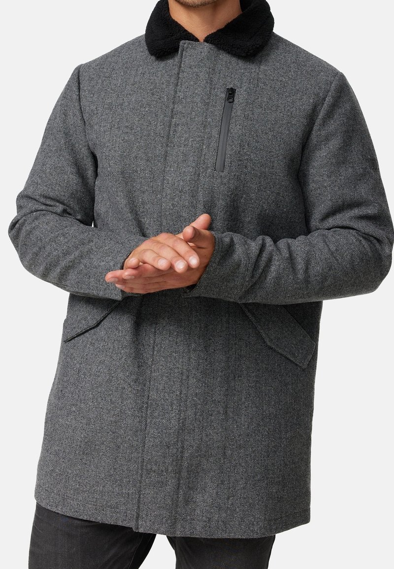 Abrigo gris de espiga con un cuello de piel sintética negro, bolsillo delantero con cremallera y bolsillos laterales. Tejido grueso con un diseño estructurado y un corte holgado.