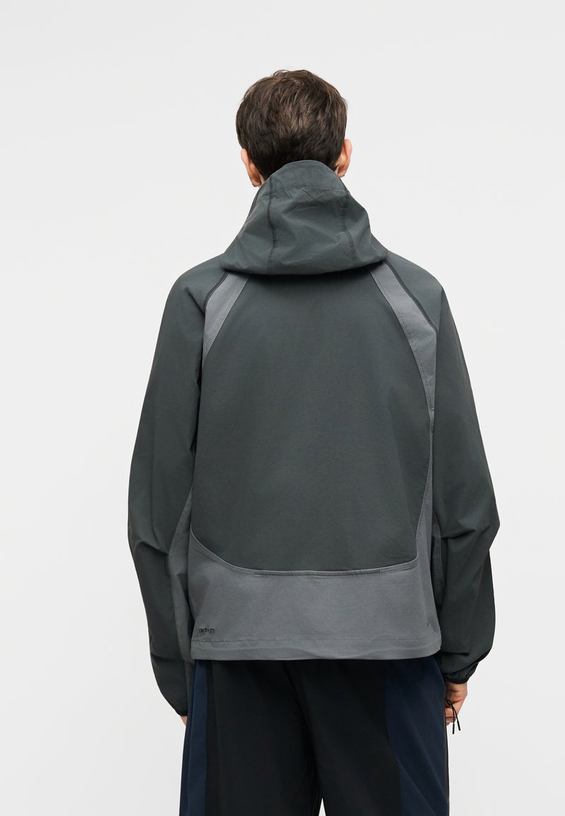 Person trägt eine dunkelgraue Kapuzenjacke mit hellgrauen Einsätzen und steht mit dem Rücken zur Kamera vor einem schlichten Hintergrund.