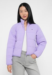 Svijetlo ljubičasti puffer jakna s visokim ovratnikom, s dijagonalnim kvadratnim šavovima i malim logom na lijevoj prsnoj strani. Vidljivo zatvaranje na zip.