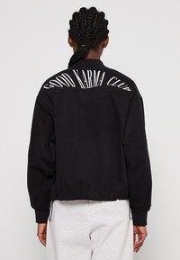 Czarna kurtka typu bomber z prążkowanym kołnierzem, elastycznymi mankietami i dołem. Na plecach znajduje się napis „GOOD KARMA CLUB” w grubych białych literach.