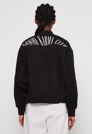 Svart bomberjakke med ribbestrikket krage, elastiske mansjetter og kant. På baksiden står det "GOOD KARMA CLUB" med fete, hvite bokstaver.