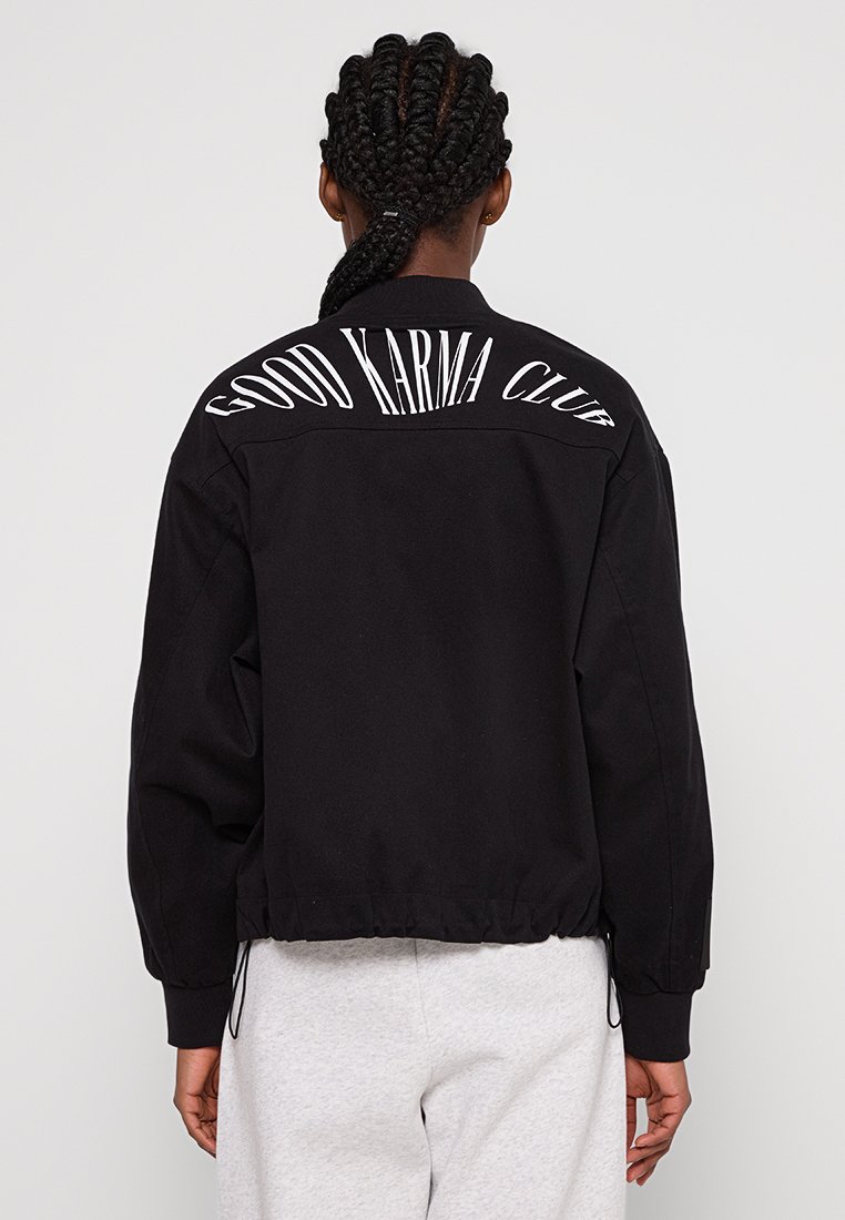 Czarna kurtka typu bomber z prążkowanym kołnierzem, elastycznymi mankietami i dołem. Na plecach znajduje się napis „GOOD KARMA CLUB” w grubych białych literach.
