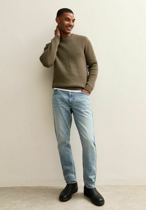 Licht olijfgroene gebreide trui, lichtblauwe jeans met een rechte snit, zwarte laarzen. Model is afgebeeld in een ontspannen, staande houding tegen een neutrale achtergrond.