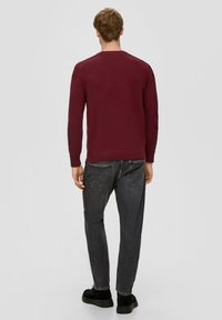 Burgunderfarbener Pullover mit gerippter Struktur, Rundhalsausschnitt und langen Ärmeln, kombiniert mit schwarzen Jeans mit einem subtilen Waschungseffekt und normalen Taschen.