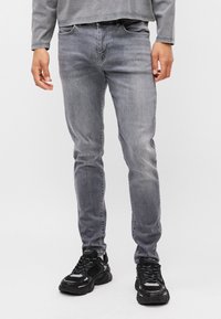 Szürke farmernadrág, amely karcsúsított szabású és finoman kopott, fekete sneakers-szel párosítva, amelyek réteges, texturált dizájnnal rendelkeznek.