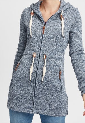 Femme portant un sweat à capuche bleu chiné avec fermeture éclair, cordons blancs et poches zippées, associé à un jean bleu.