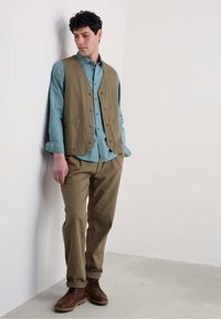 Beige vest over een lichtblauw gestreept shirt, gecombineerd met kaki broek en bruin veterschoenen. Minimalistisch ontwerp met knopen en zakken.