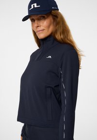 Giacca a zip blu navy con collo alto, con logo sul petto e testo riflettente lungo le maniche. Accompagnata da un cappellino abbinato.