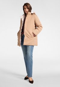 Béžový puffrový kabát s bílou podšívkou, se zasouvací kapucí, bočními kapsami a zlatými cvočky. Kombinováno s modrými džíny a hnědými plochými botami.