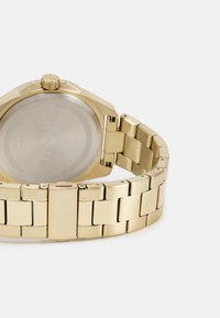 Reloj de metal tono dorado con una cara rectangular, brazalete de eslabones y caja octagonal. Presenta una corona en la parte superior y un respaldo metálico liso.