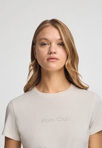Beżowy t-shirt z krótkim rękawem wykonany z bawełny, z okrągłym dekoltem i nadrukiem "Polo Club" w kropkowanym wzorze z przodu. Gładka tekstura.
