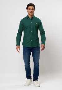 Zöld gombos ing, hosszú ujjakkal, kicsi logóval a mellrészen. Kék farmernadrággal és fehér sportcipővel párosítva.