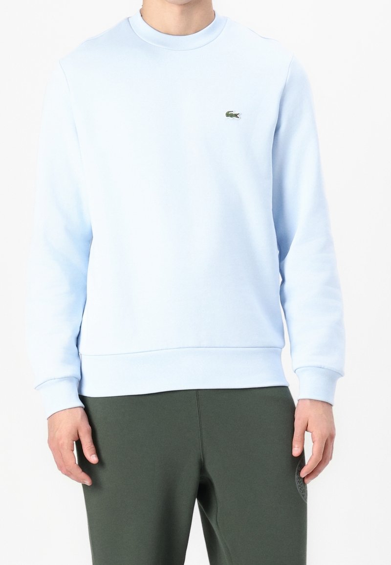 Ljusblå crewneck sweatshirt i bomullsblandning, med en liten grön logotyp på vänster bröst och ribbade ärmslut och fåll.