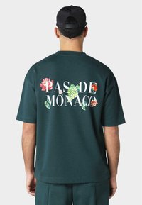 Mørkegrønn oversized T-skjorte med blomsterprint og teksten "PAS DE MONACO" i hvitt. Glatt stoff og korte ermer.