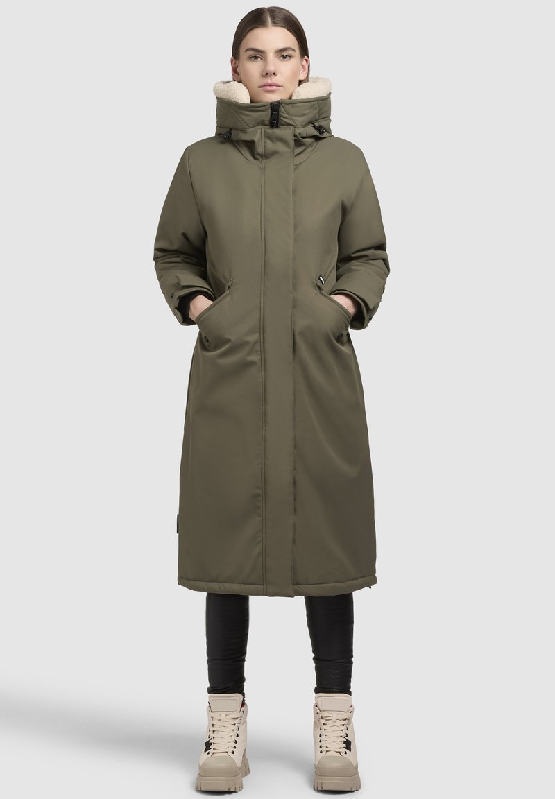 Olivi vihreä pitkä parka, jossa on fleecevuorattu huppu, kaksi sivutaskua ja edessä vetoketju. Materiaali on sileä ja hieman kiiltävä.