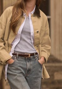 Beige kangaskovakaitaliha kangaskovakaitaliha, jossa on samettikaulus, kerrostettuna harmaan t-paidan ja valkoisen napitettavan paidan päälle, yhdistettynä vaaleansinisiin farkkuihin ja ruskeaan vyöhön.