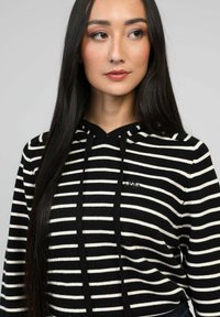 Hoodie rayé avec des bandes horizontales noires et blanches, capuche avec cordons de serrage et un petit logo sur le devant. Tissu tricoté doux.