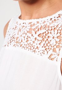 Robe blanche présentant un col en dentelle à motifs floraux et un corps en tissu lisse. Le contraste de texture entre la dentelle et le tissu est marqué.