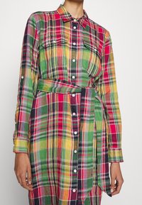 Langärmliges Karohemdkleid mit Knopfverschluss, Brusttaschen und Stoffgürtel in grünen, roten, gelben und schwarzen Tönen an einer stehenden Person.