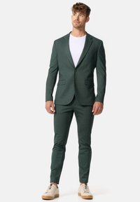 Ciemnozielony garnitur wykonany z teksturowanej tkaniny, z dopasowanym krojem, klapami z wycięciem i dwoma przednimi kieszeniami, zestawiony z gładką białą koszulą i beżowymi butami.