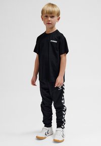 Svart bomull t-shirt med vit "HUMMEL" logotyp, ihopparad med svarta joggers som har vita chevronränder och vita skor med gummisålar.