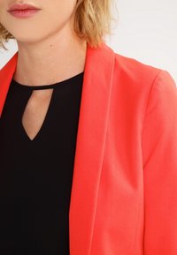 Korallblazer med en skräddarsydd design, med slag och slät textur, bärs över en svart topp med detaljer i nyckelhålsform.