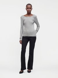 Femme debout portant un haut gris ajusté à manches longues, un pantalon noir évasé et des chaussures noires à bout pointu sur un fond blanc uni.
