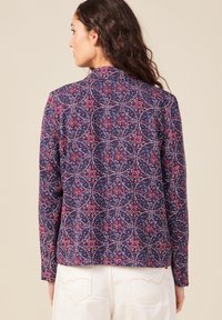 Camicia a maniche lunghe in tessuto blu navy con un motivo geometrico colorato e dettagli rossi e bianchi. Taglio dritto con colletto alto.