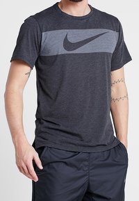Camiseta deportiva de color gris oscuro con mangas cortas, que presenta un patrón de rayas horizontales en blanco y negro y un prominente logo negro de Nike.