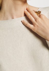 Beige strukturierter Pullover mit weitem Halsausschnitt, kombiniert mit einem goldenen abstrakten Ring, der an der rechten Hand getragen wird und das minimalistische Design betont.