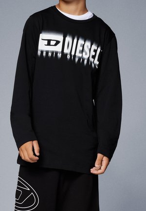 Kind draagt een zwart shirt met lange mouwen met witte "DIESEL"-logo en bijpassende zwarte shorts met wit grafisch ontwerp op het linkerbeen.