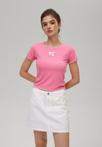 Roosa ribaga lühikeste varrukatega topp, millel on lainelised servad ja valge lips detailina rinnal, kombineeritud valge denim mini seelikuga.