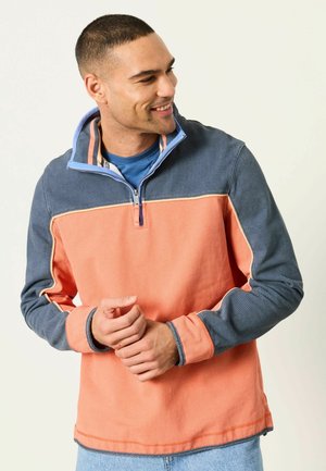 Junger Mann, der lächelt und einen halben Reißverschluss-Sweatshirt mit korallenfarbenem Körper und blauen Ärmeln trägt, vor einem schlichten hellen Hintergrund steht.