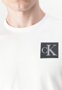 Vit bomull t-shirt med en svart rektangulär logotyp med stiliserat "cK"-mönster på vänster bröstområde. Rund halsdesign.