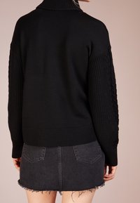 Pull noir en tricot avec un col montant, présentant des détails en côtes et en torsades sur les manches, associé à une jupe en denim noir à ourlet effiloché.