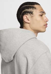 Siva jopa z velikim, sproščenim kapuco. Tkanina je mehka z gladko teksturo. Značilnosti vključujejo preprost dizajn in vidne šive.