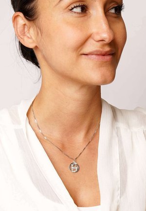 Frau mit einer silbernen Halskette und einem runden Anhänger, der ein kleines Tierdesign zeigt, gekleidet in einer weißen Bluse.