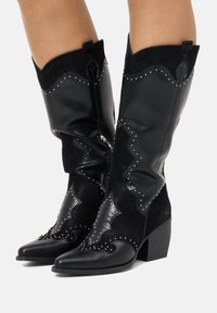 Botas de cowboy negras con punta afilada, que presentan una combinación de cuero liso y ante, un intrincado diseño acanalado y tachuelas plateadas.