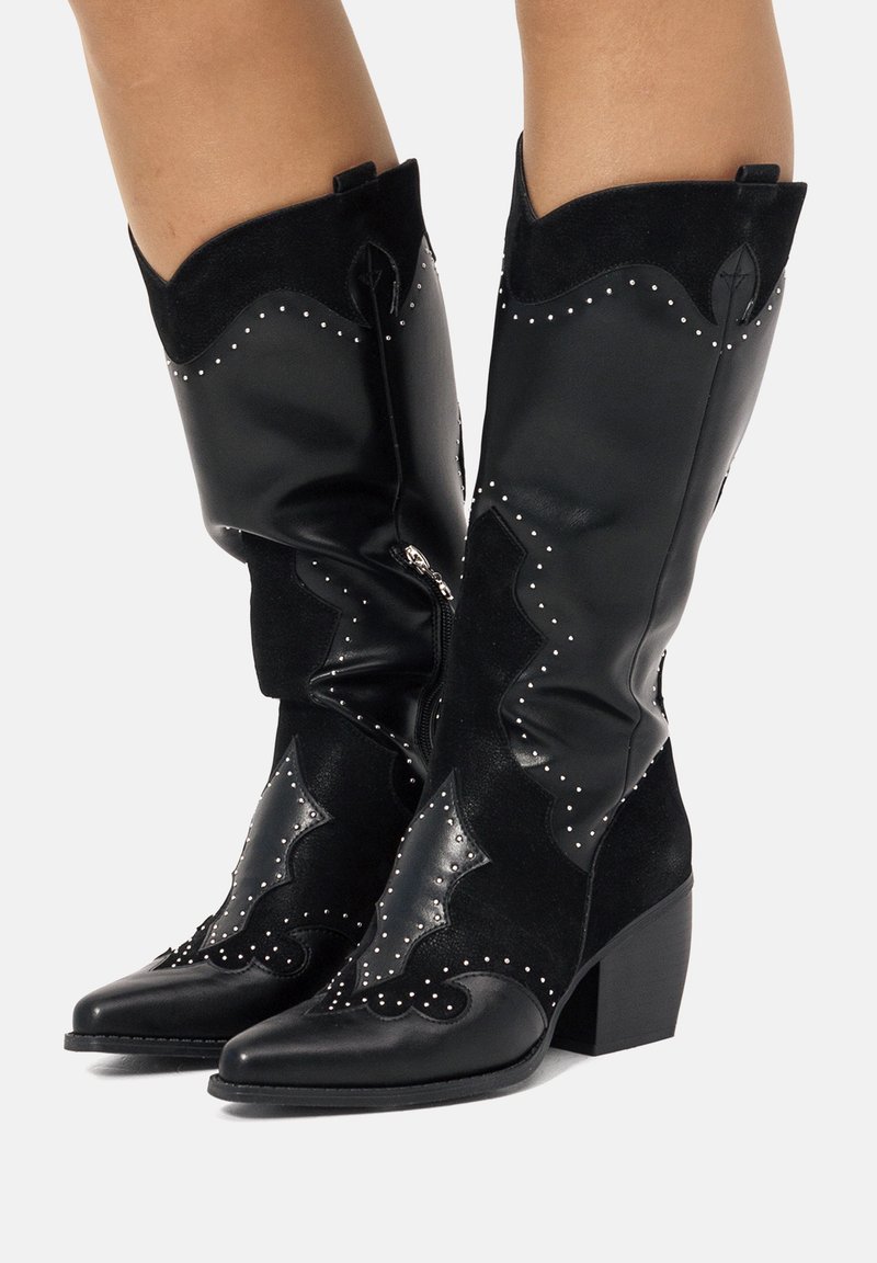 Botas de cowboy negras con punta afilada, que presentan una combinación de cuero liso y ante, un intrincado diseño acanalado y tachuelas plateadas.