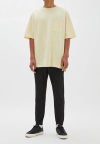 Personne portant un T-shirt beige clair surdimensionné avec une poche poitrine, un pantalon noir, des chaussettes blanches et des baskets noires, debout devant un fond uni.